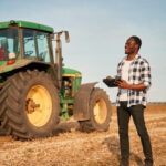Jeune agriculteur noir souriant, debout dans un champ, tenant une tablette numérique avec un grand tracteur vert en arrière-plan