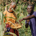 Deux agriculteurs (homme et femme) souriants dans un champ, l'homme présentant des produits agricoles dans un panier, symbolisant la récolte ou la production.