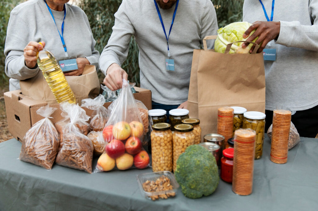 Bénévoles distribuant des denrées alimentaires variées (légumes, conserves, huile, fruits secs) lors d'une action de soutien alimentaire.