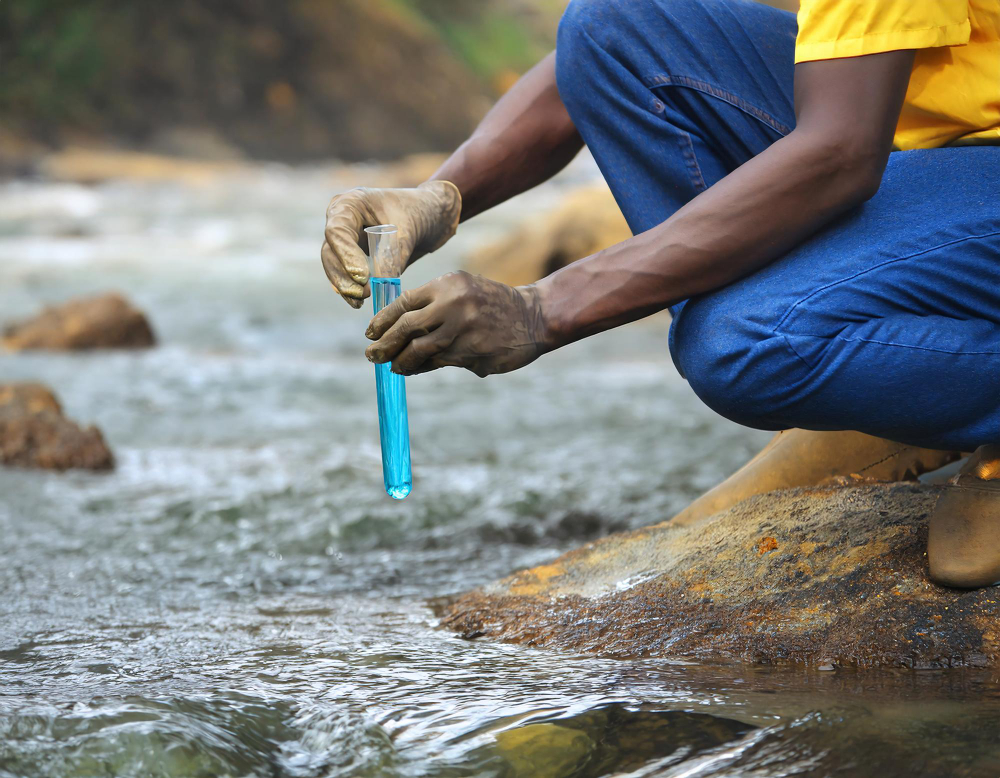 Personne effectuant un prélèvement d'eau dans un cours d'eau à l'aide d'une éprouvette bleue pour analyse