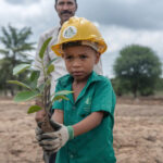 Enfant avec un casque de protection et des gants tenant une jeune pousse d'arbre, prêt à planter, avec un adulte en arrière-plan